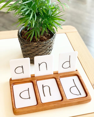 Three white dry erase flash cards with letters and words in black marker in wooden holder on white table next to plant