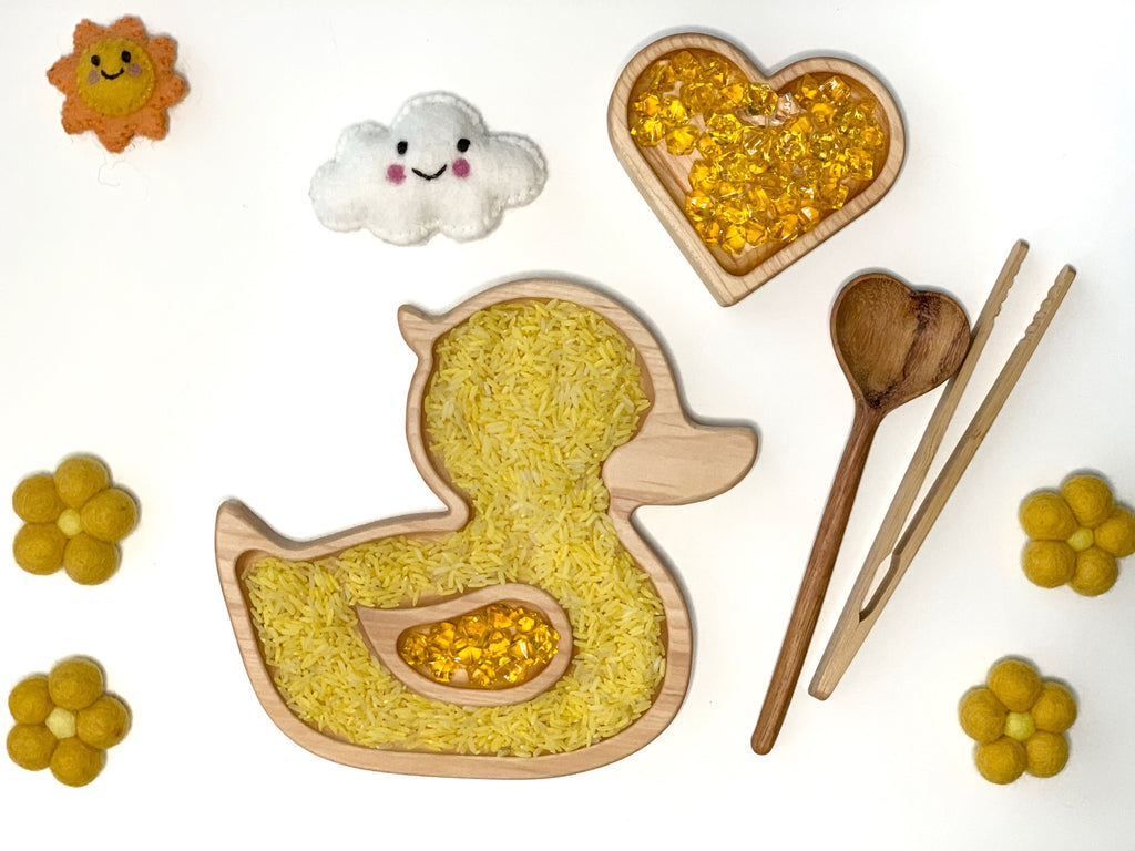 Wooden duck-shaped sensory tray filled with yellow rice and gems next to heart tray and wooden utensils on white background