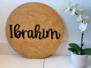 Round wooden name sign with black cursive letters spelling Ibrahim on natural wood background next to white orchid plant