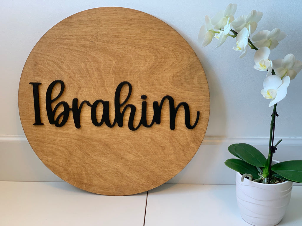 Round wooden name sign with black cursive letters spelling Ibrahim on natural wood background next to white orchid plant