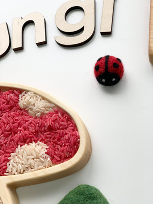 Small handmade red and black felt ladybug with white eyes on white background near wooden letters and colored rice in wood tray