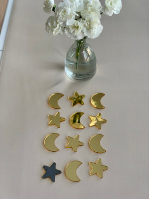 Set of twelve glossy gold acrylic moon and star shapes arranged in rows on beige surface with white flowers in glass vase