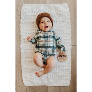 Baby lying on white textured blanket wearing plaid onesie and brown knit hat holding seven months wooden milestone disc