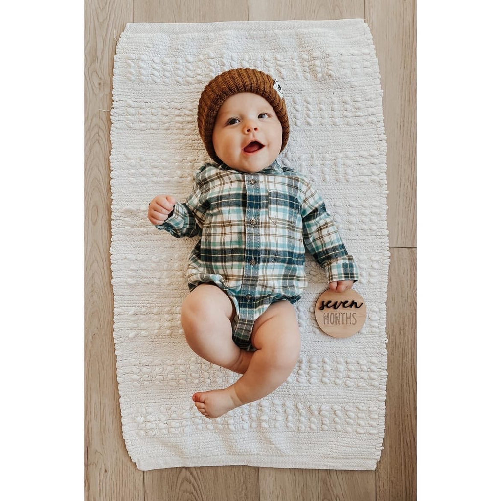 Baby lying on white textured blanket wearing plaid onesie and brown knit hat holding seven months wooden milestone disc