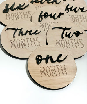 Round wooden monthly milestone discs with black cursive numbers and engraved text on light wood background