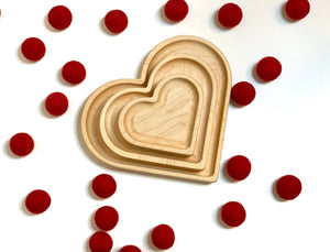 Three natural wooden nesting heart plates arranged by size surrounded by scattered red felt balls on white background