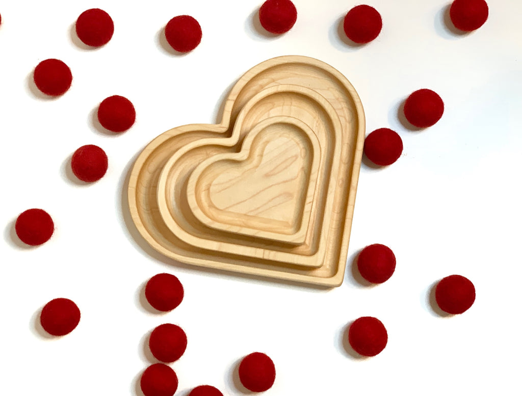 Three natural wooden nesting heart plates arranged by size surrounded by scattered red felt balls on white background
