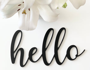 Black wooden word cutout reading hello in cursive script with white lily flower on white background
