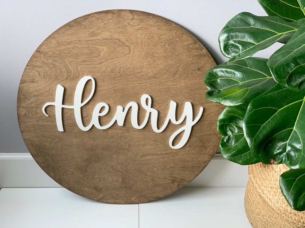 3D round wooden nursery sign with white cursive name lettering on natural wood grain background next to green plant