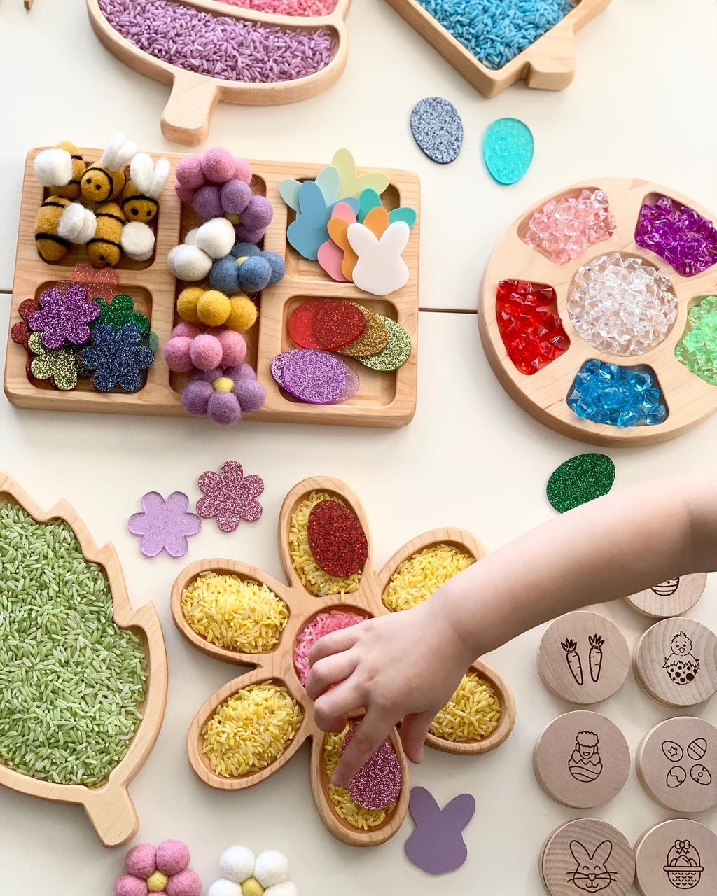 Wooden trays filled with colorful sensory play acrylic shapes, pom poms, and rainbow rice being arranged by child's hand