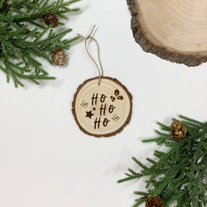 Round wood slice Christmas ornament with Ho Ho Ho text and holly design on white background with pine and pinecones