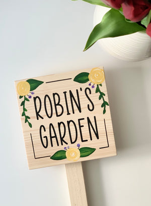 Small square natural wooden garden sign with black letters and yellow floral illustrations on stake white background