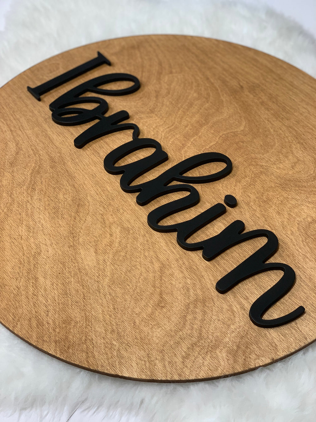 Round wooden name sign with black 3D cursive letters on natural wood grain background
