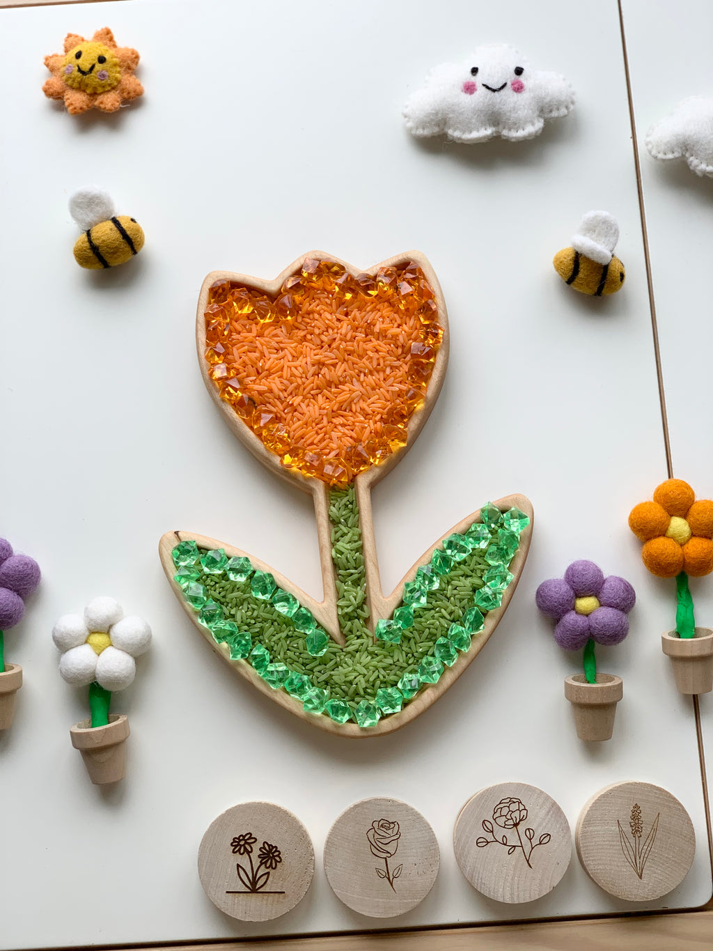 Wooden tulip-shaped sensory tray filled with orange rice and green gems surrounded by felt bees and flowers on white surface