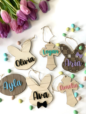 Custom wooden Easter basket name tags in bunny and cross shapes with pastel colored names and twine hangers on white background