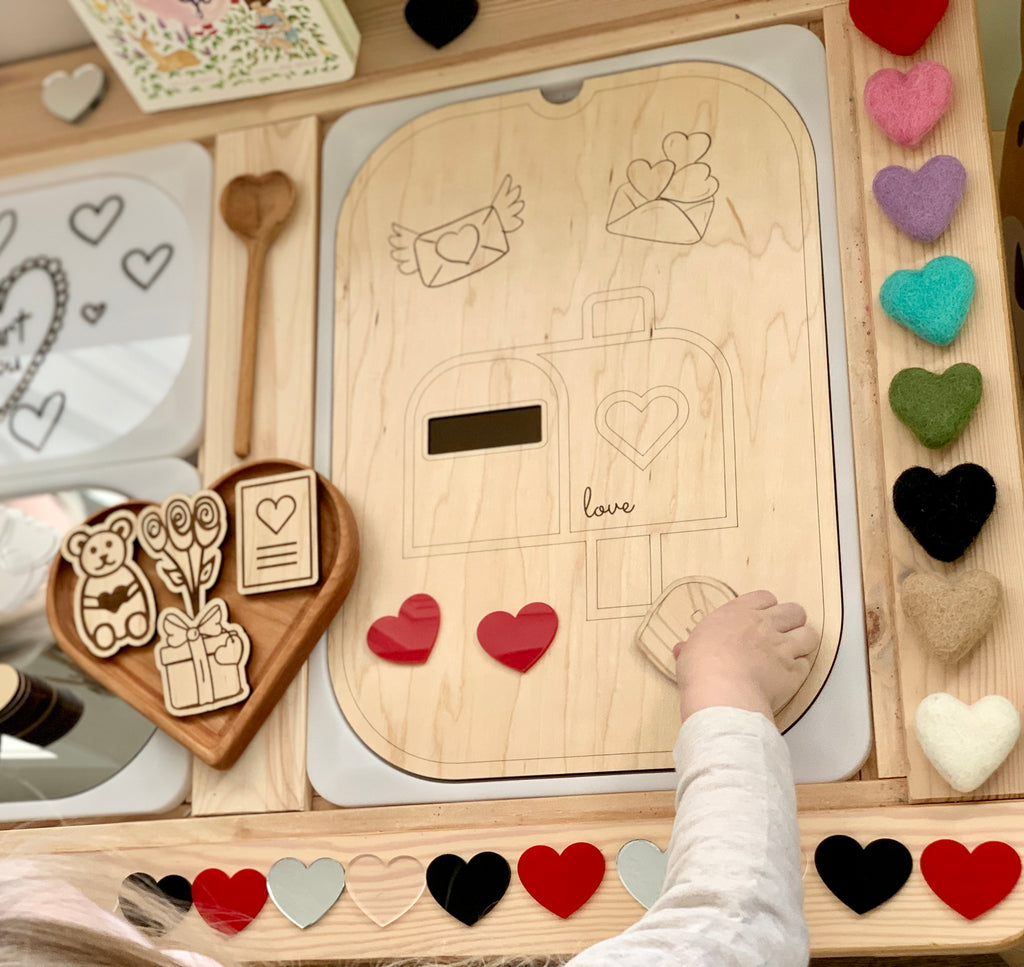 Child’s hand placing heart-shaped acrylic pieces on wooden mailbox sensory play board with felt and wooden hearts around edges