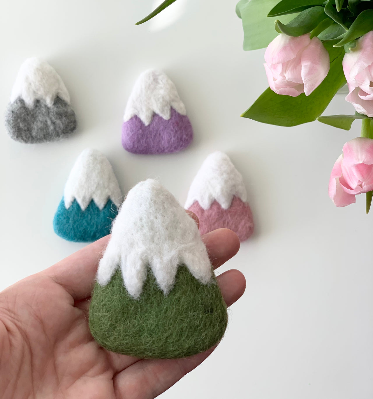 Handmade felt mountain in green with white snowy peak held in hand with other colorful felt mountains and pink flowers nearby