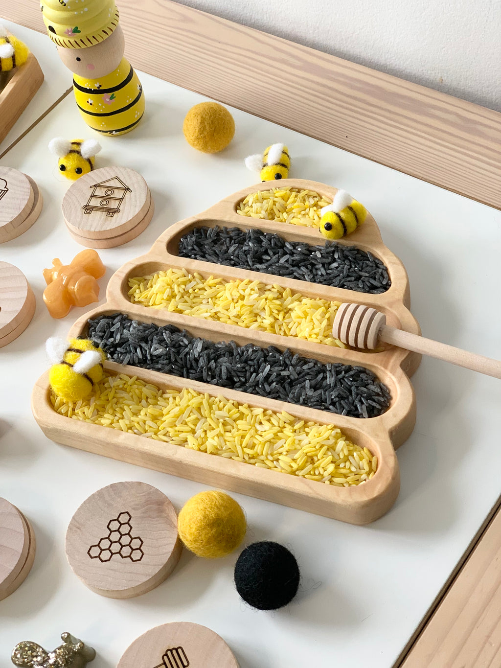 Beehive-shaped wooden sensory tray filled with yellow and black rice, wooden honey dipper, and felt bee toys on white surface