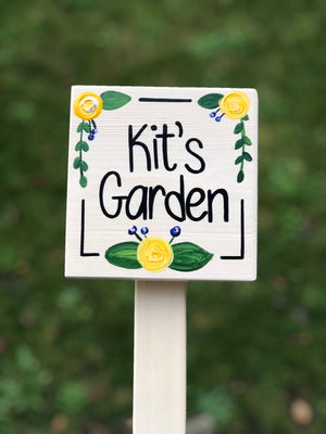 Small square wooden garden sign painted white with black lettering and yellow floral accents on stake background blurred green grass