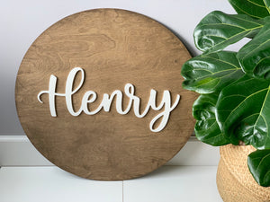 Large round wooden name sign stained brown with white cursive letters spelling Henry near green potted plant