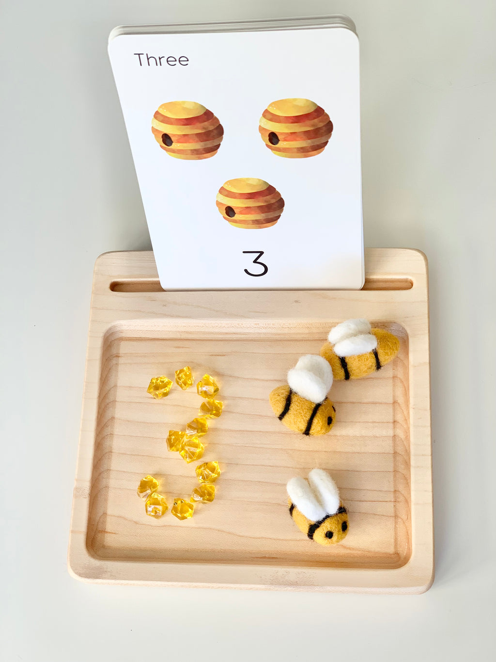 Light wooden sensory tray with three yellow gem beads and three small felt bee figures with a number three flash card
