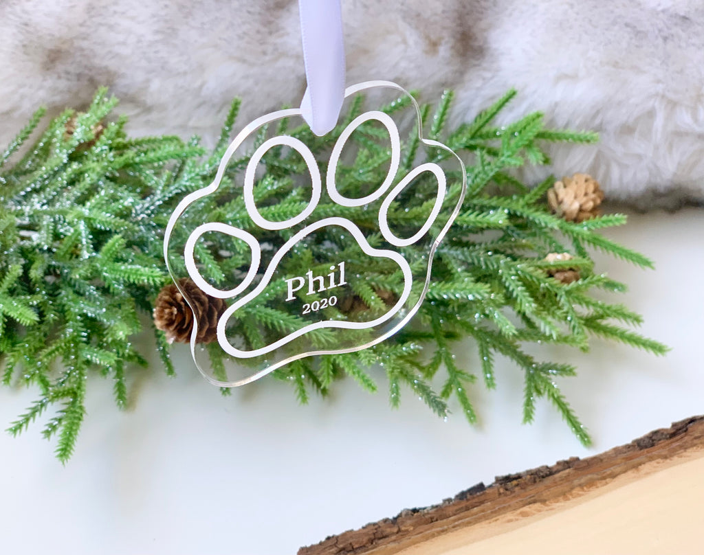 Clear acrylic dog paw print ornament engraved with name Phil and year 2020 hanging on white ribbon over green pine branch