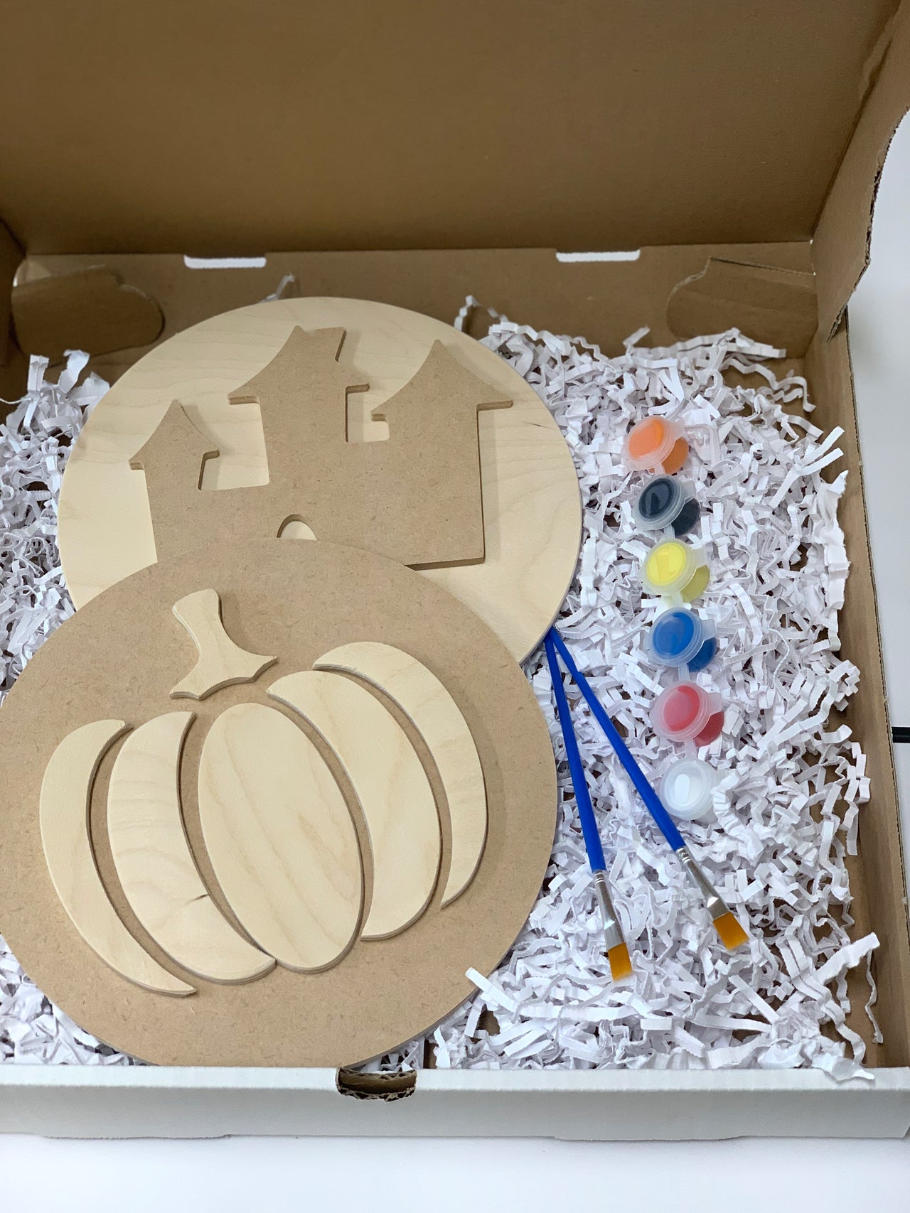DIY Halloween sign kit with unfinished wooden pumpkin and haunted house shapes, paint pots, and brushes in cardboard box