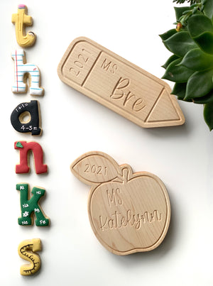 Two personalized wooden teacher name plates shaped like a pencil and an apple on white background with colorful alphabet magnets and green plant