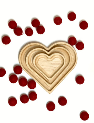 Natural wooden nesting heart plates arranged by size on white surface with scattered small red felt balls