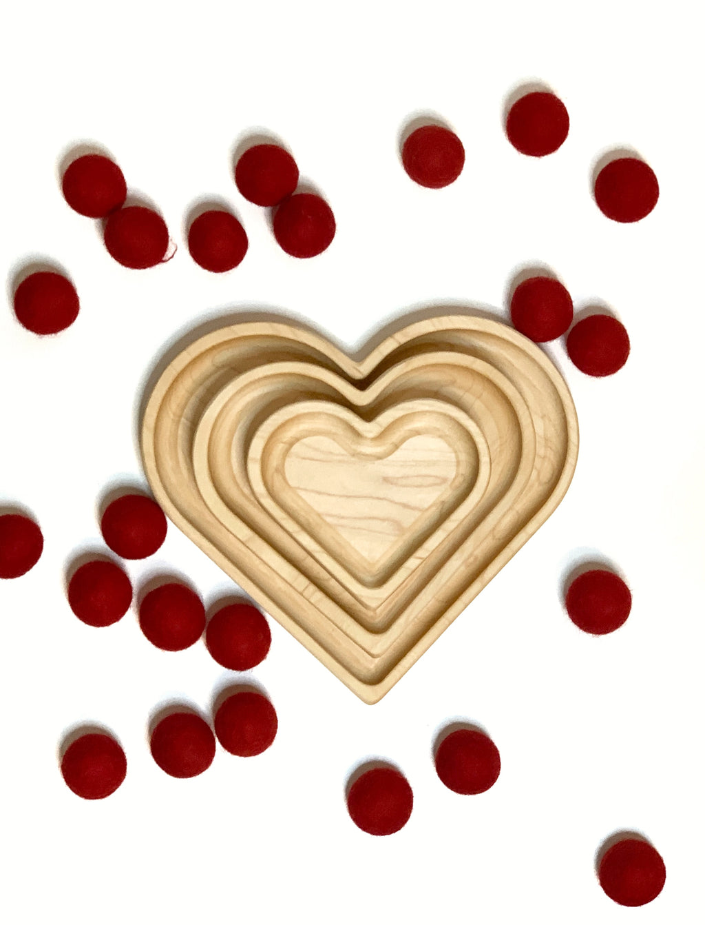Natural wooden nesting heart plates arranged by size on white surface with scattered small red felt balls