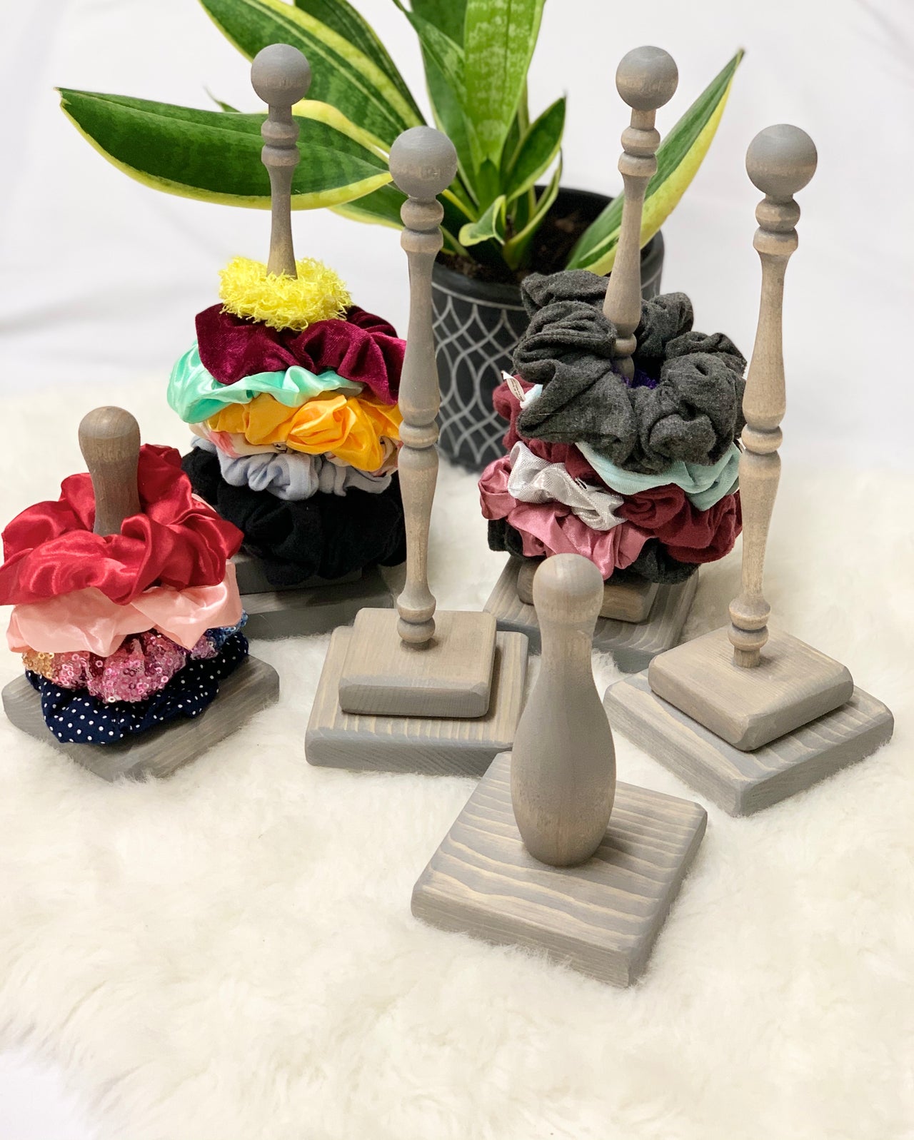 Natural wooden scrunchie holders with square bases holding colorful fabric scrunchies on white fuzzy surface with green plant background