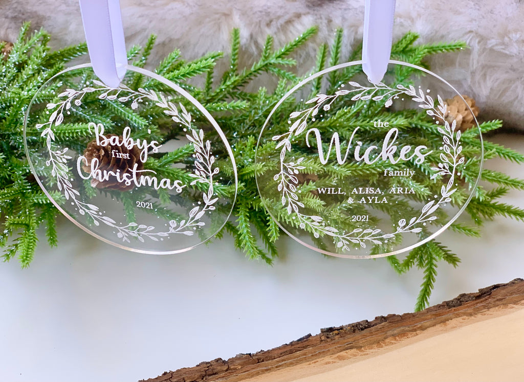 Two clear acrylic Christmas ornaments engraved with white floral wreath designs and personalized text hanging on greenery background