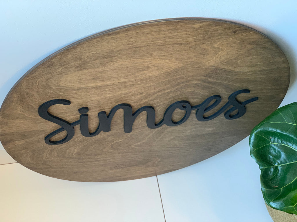Oval wooden 3D round name sign stained brown with black cursive letters on white surface near green plant leaf