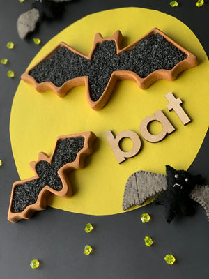 Two wooden bat-shaped sensory trays filled with black rice on yellow and black background with wooden letters spelling bat