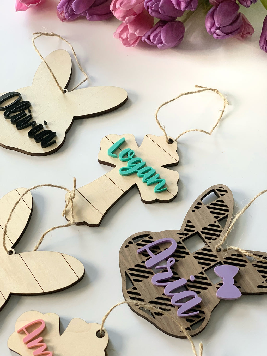 Wooden Easter basket name tags in bunny and cross shapes with colorful acrylic names and twine hangers on white surface with purple flowers
