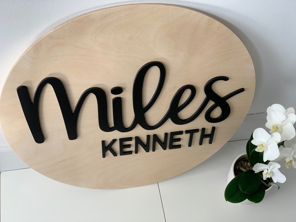 Oval natural wood 3D round name sign with black cursive and block letters on white surface near white orchid plant