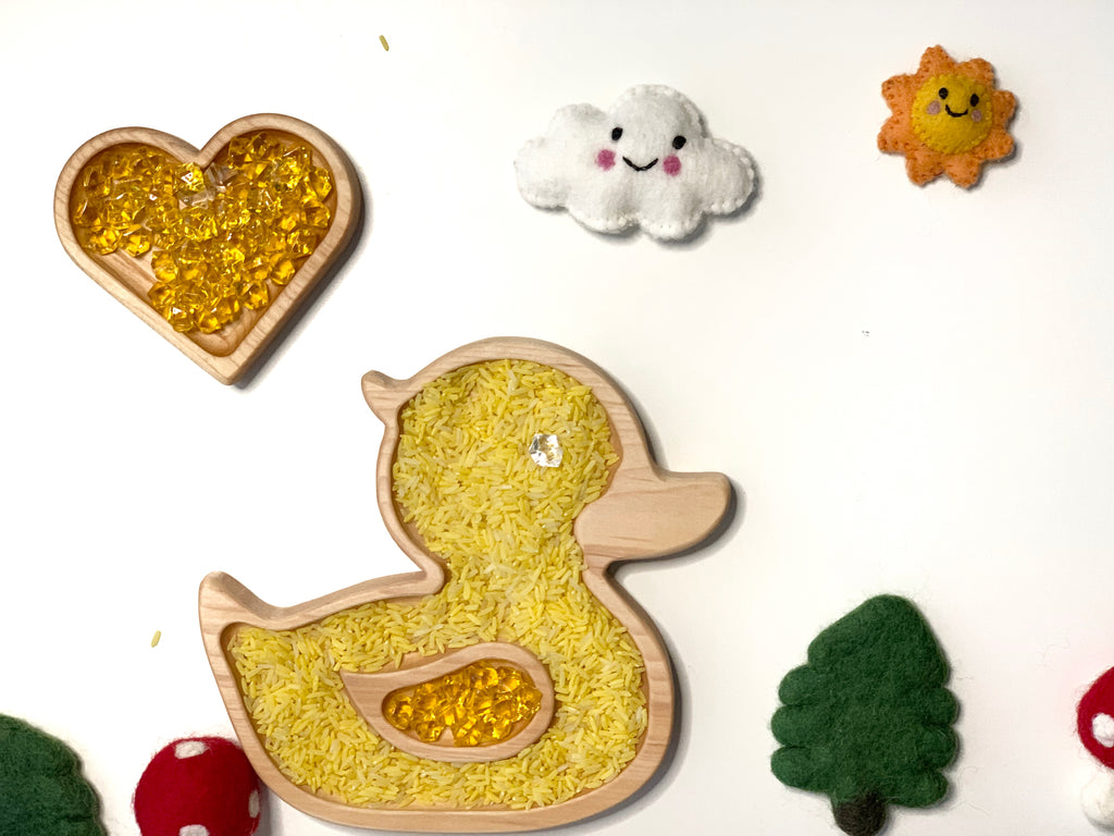 Natural wooden duck-shaped sensory tray filled with yellow rice and beads on white background with felt decorations