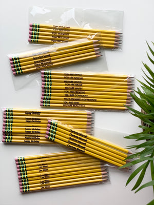 Sets of yellow pre-sharpened affirmation and personalized pencils with pink erasers in clear plastic packaging on white surface