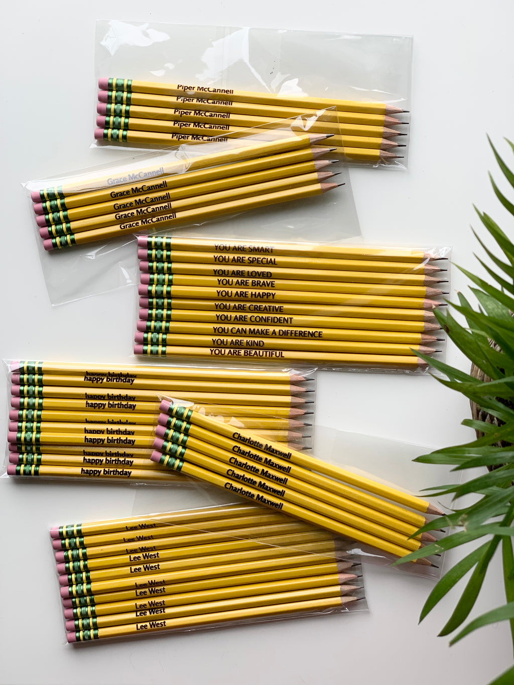 Sets of yellow pre-sharpened affirmation and personalized pencils with pink erasers in clear plastic packaging on white surface