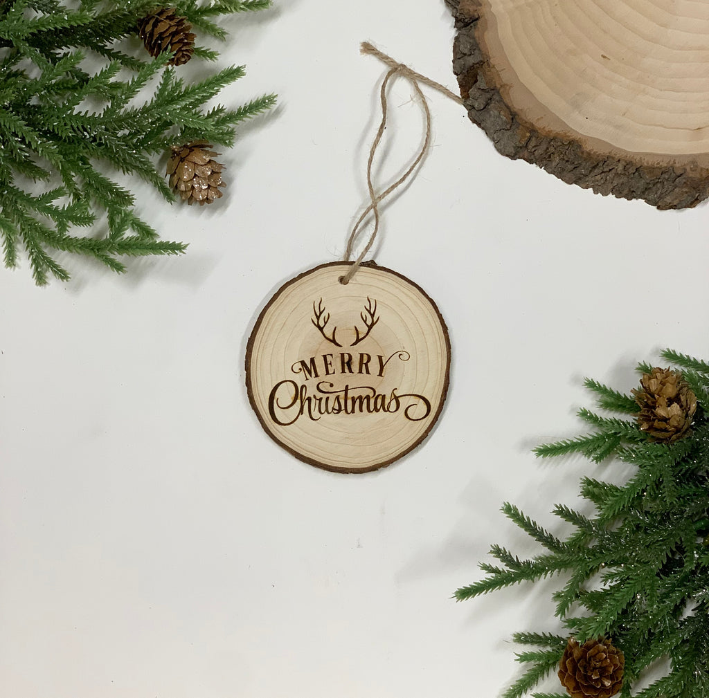 Round wood slice Christmas ornament with burned “Merry Christmas” and antler design on white background with greenery and pinecones
