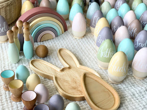 Hollow wooden Easter eggs in pastel colors with white names, natural wood bunny tray, wooden egg cups, and bowling pins on fabric surface
