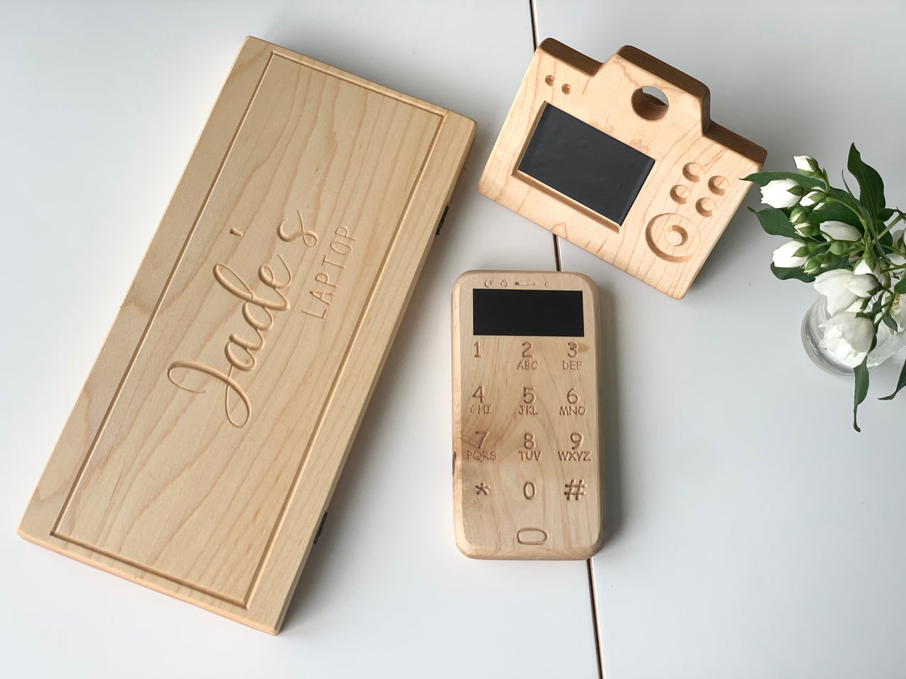 Set of natural wooden toy laptop, camera with engraved buttons and screen, and a phone on white surface with green plant
