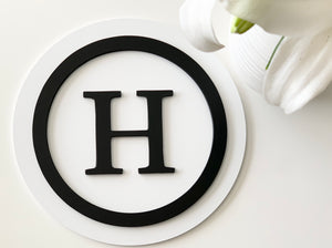 3D monogram round sign with black letter H and circle on white layered MDF background near white flower