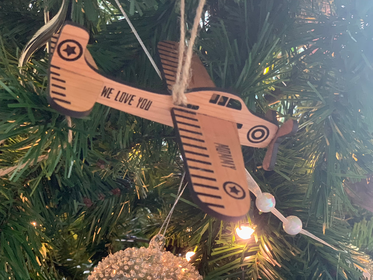 Wooden airplane Christmas ornament with custom engraved text hanging on green tree with lights and decorations