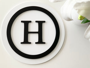 Round 3D monogram sign with black letter H and circle on layered white wooden background with flowers nearby