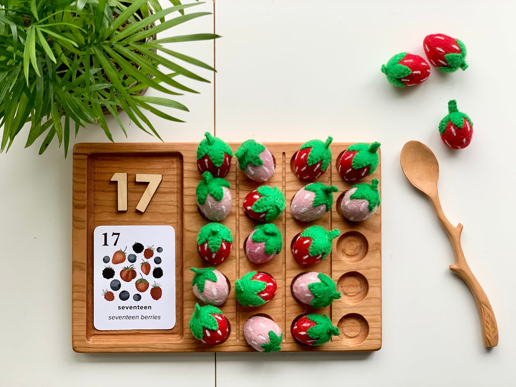 Natural wooden twig style spoon beside wooden counting tray with felt strawberries and number blocks on white surface