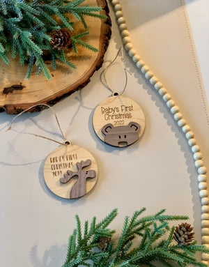 Two wooden baby’s first Christmas ornaments with moose and bear designs, natural greenery and wood slice background