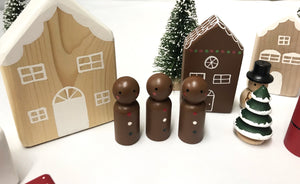 Small handmade Christmas peg dolls painted brown with festive trees and wooden house decorations on white surface