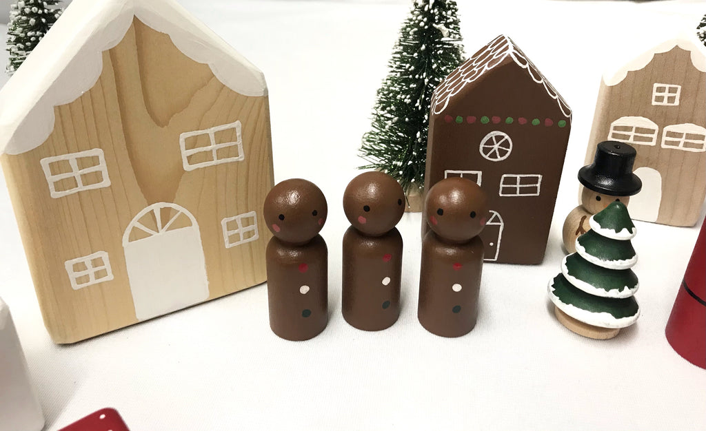Small handmade Christmas peg dolls painted brown with festive trees and wooden house decorations on white surface