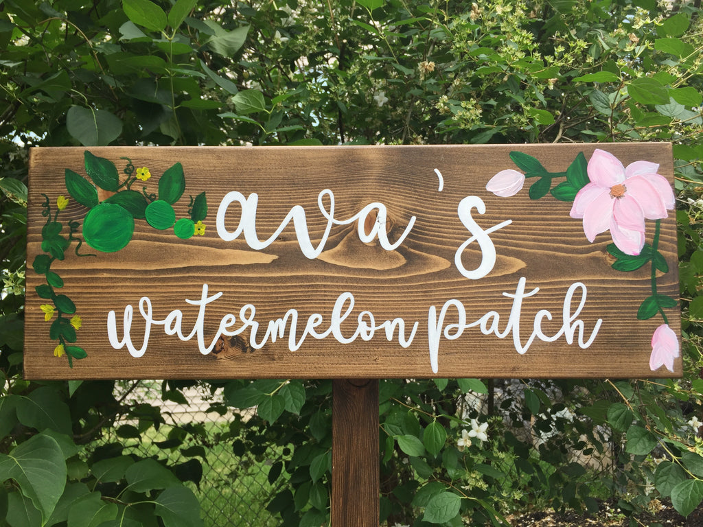 Large personalized wooden garden sign with white cursive letters and green and pink floral illustrations on natural wood background